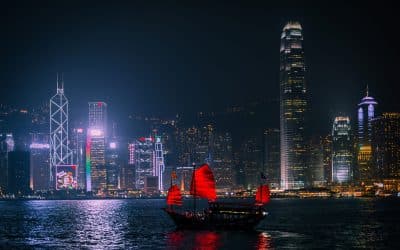 Hong Kong Island skyscrapers lit up at night behind Victoria Harbour with a red-sailed junk boat in the foreground