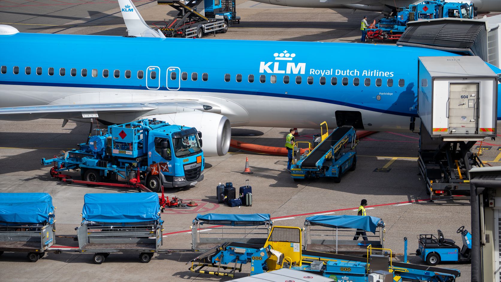 Overhead view of a blue KLM Royal Dutch Airlines jet at a gate, with fuel trucks, baggage loaders, covered carts and ground crew working around it