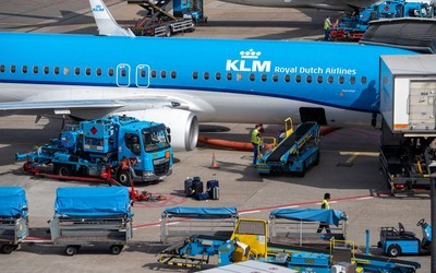Overhead view of a blue KLM Royal Dutch Airlines jet at a gate, with fuel trucks, baggage loaders, covered carts and ground crew working around it