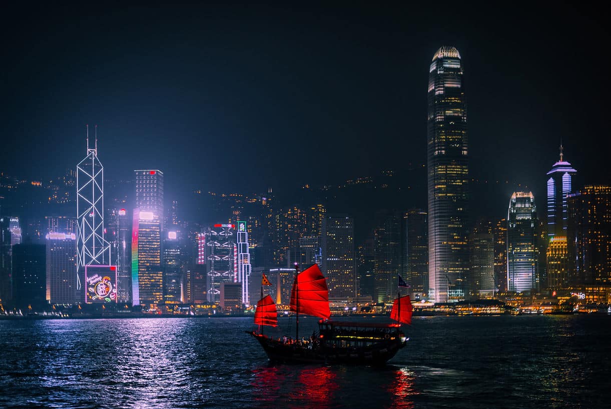 Hong Kong Island skyscrapers lit up at night behind Victoria Harbour with a red-sailed junk boat in the foreground