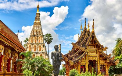 Thai temple grounds with a tall tiered chedi, a dark standing monk statue and an ornate gold-trimmed hall