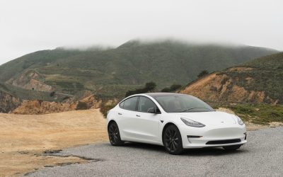 A white Tesla Model 3 sedan parked on a gravel turnout with fog-covered green mountains in the background.