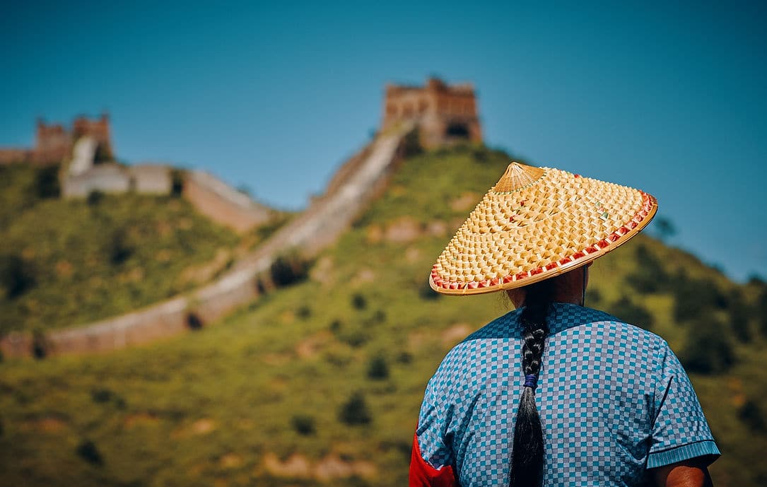 A person in a woven straw hat looking out over the Great Wall of China on a green hillside
