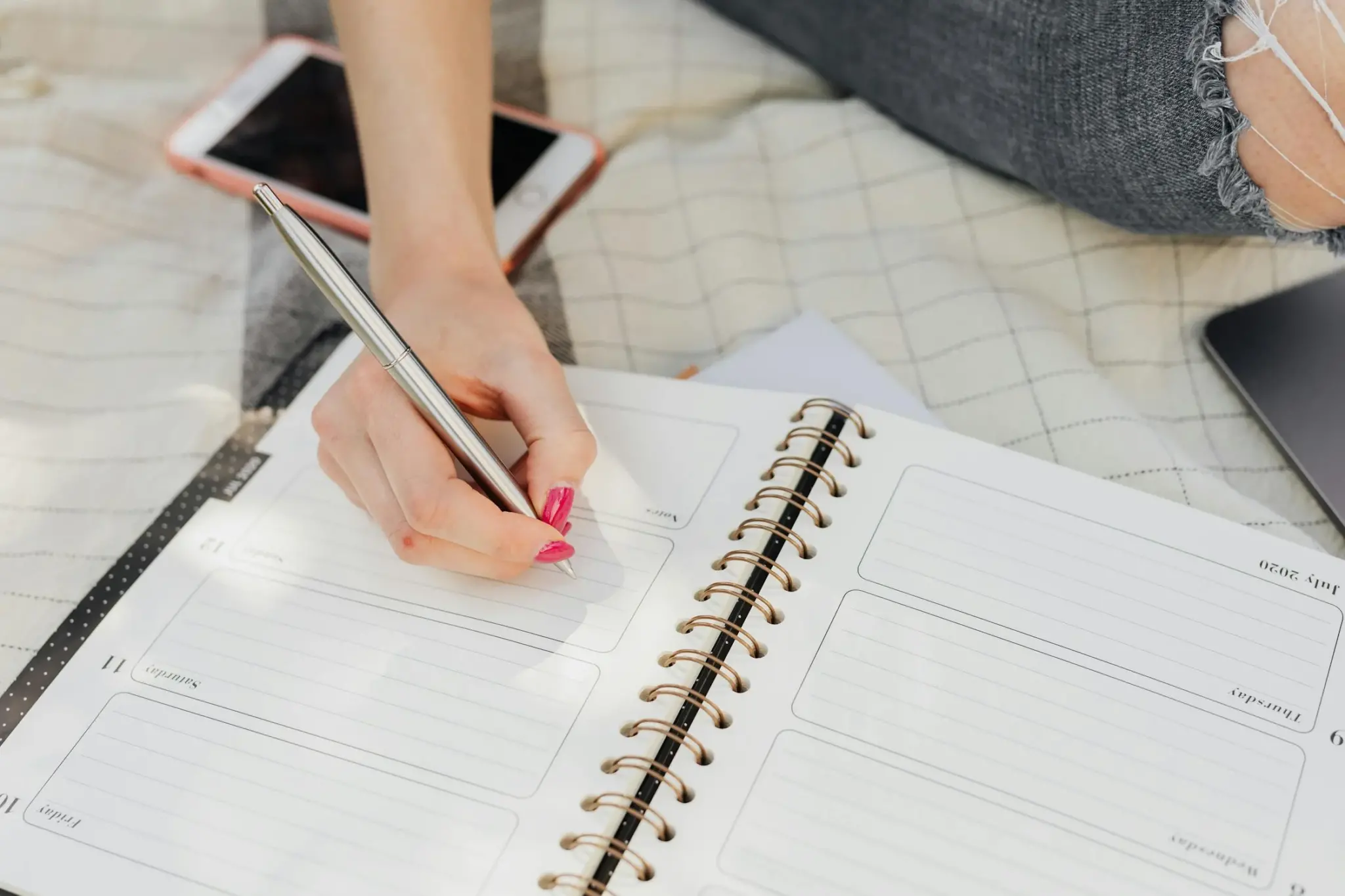 A hand with pink nail polish holds a silver pen, writing in a spiral-bound weekly planner open to a July page, with a pink phone and a dark tablet resting nearby on a light checkered blanket.
