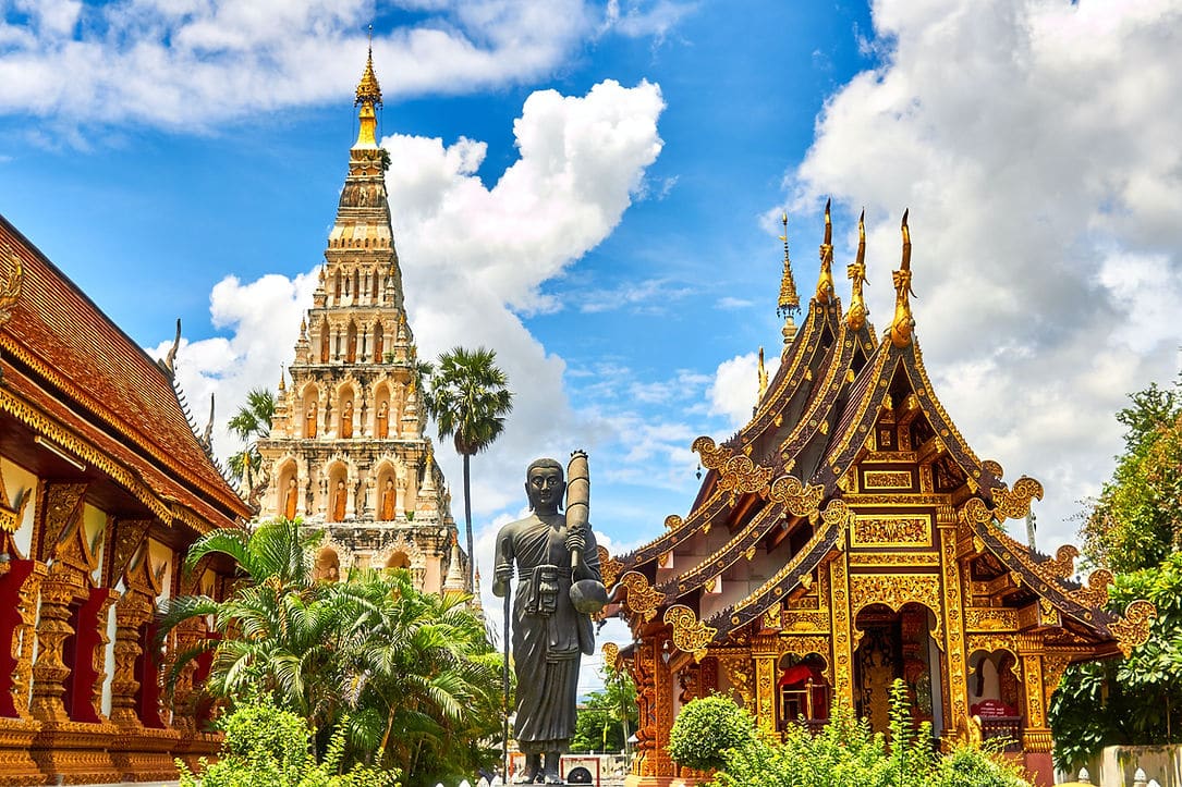 Thai temple grounds with a tall tiered chedi, a dark standing monk statue and an ornate gold-trimmed hall
