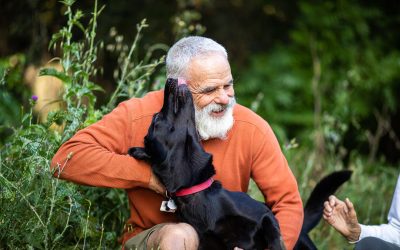 A grey-bearded older man in an orange sweater smiles as a black dog with a red collar leans up to nuzzle his face outdoors