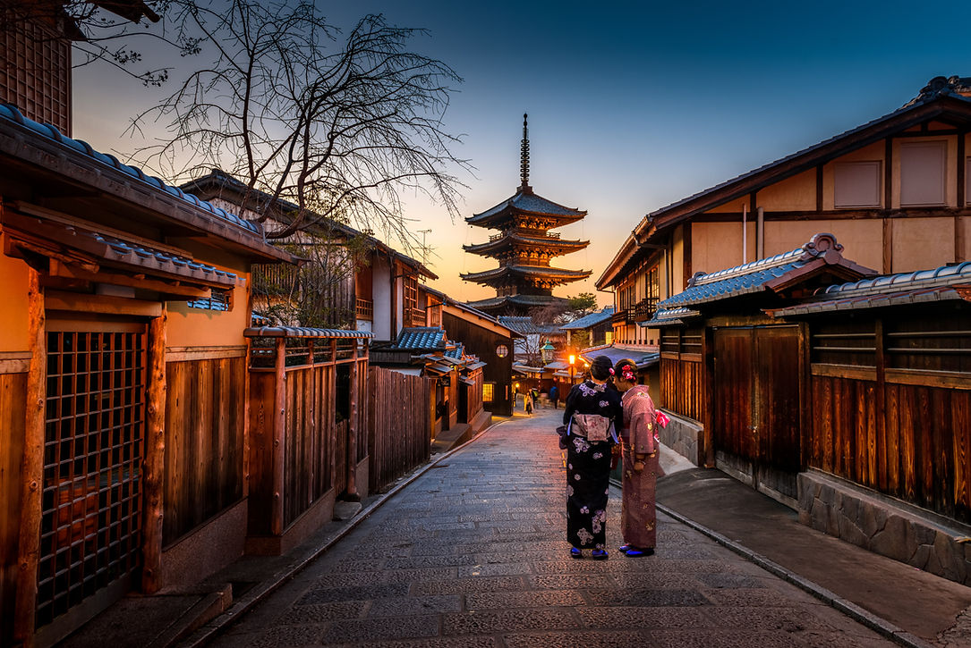 Two people in kimono walking a stone-paved Kyoto lane lined with wooden machiya houses toward a lit pagoda at dusk