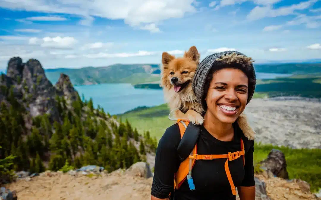 A smiling woman wearing a hiking backpack and knit beanie has a small tan long-haired dog perched on her shoulder, with a mountain lake and forest in the background.