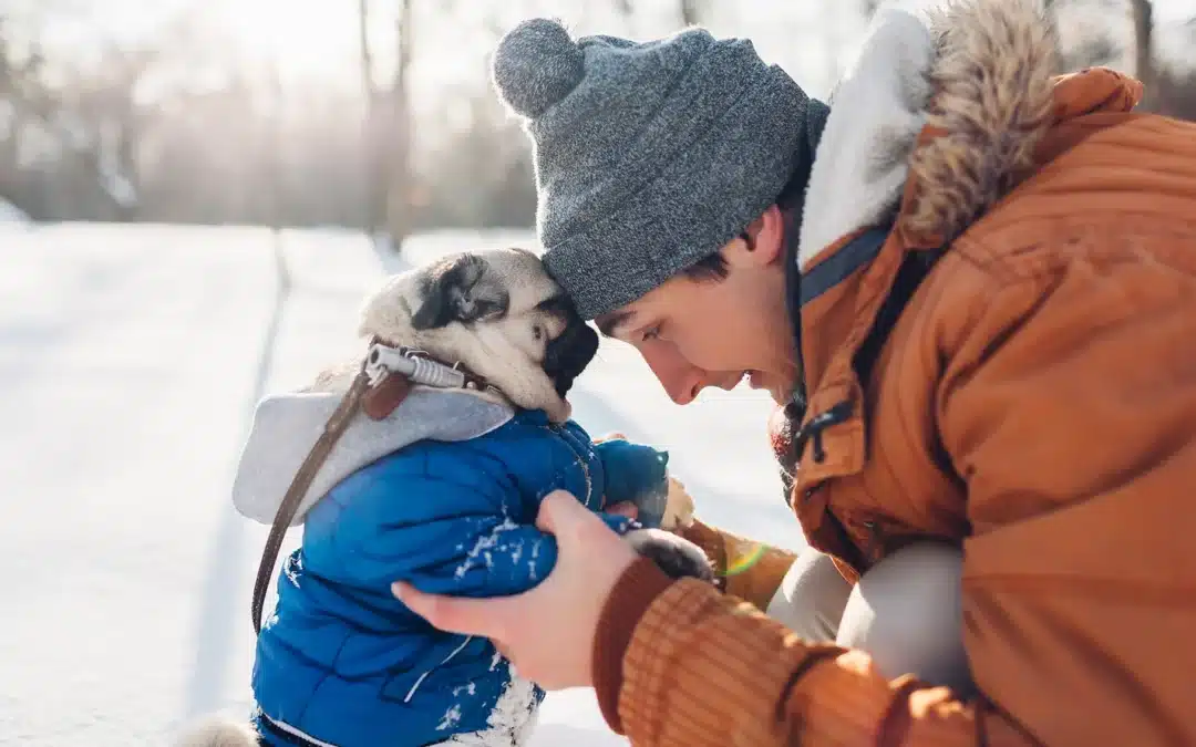 A man in an orange winter coat and gray beanie touches foreheads with a pug wearing a blue jacket, both bundled up outdoors in the snow.