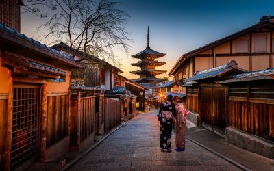 Two people in kimono walking a stone-paved Kyoto lane lined with wooden machiya houses toward a lit pagoda at dusk